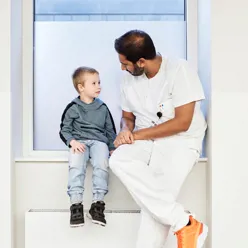 En lege i hvitt arbeidsantrekk og en gutt sitter i en vinduskarm