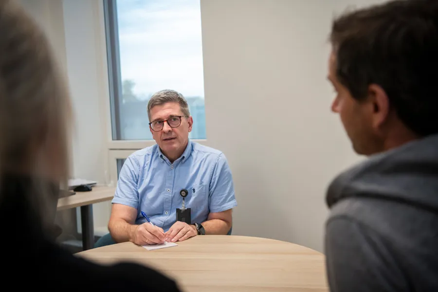 En mann som sitter ved et bord og snakker med to personer man ser bakhodet til