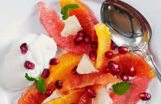 En desserttallerken med fileter av sitrusfrukt i ulike farger, toppet med granateplekjerner, mynteblader og en skje yoghurt ved siden av.