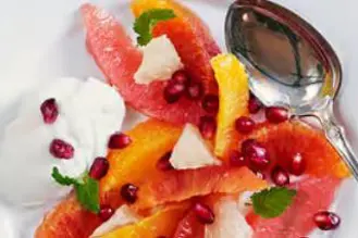 En desserttallerken med fileter av sitrusfrukt i ulike farger, toppet med granateplekjerner, mynteblader og en skje yoghurt ved siden av.