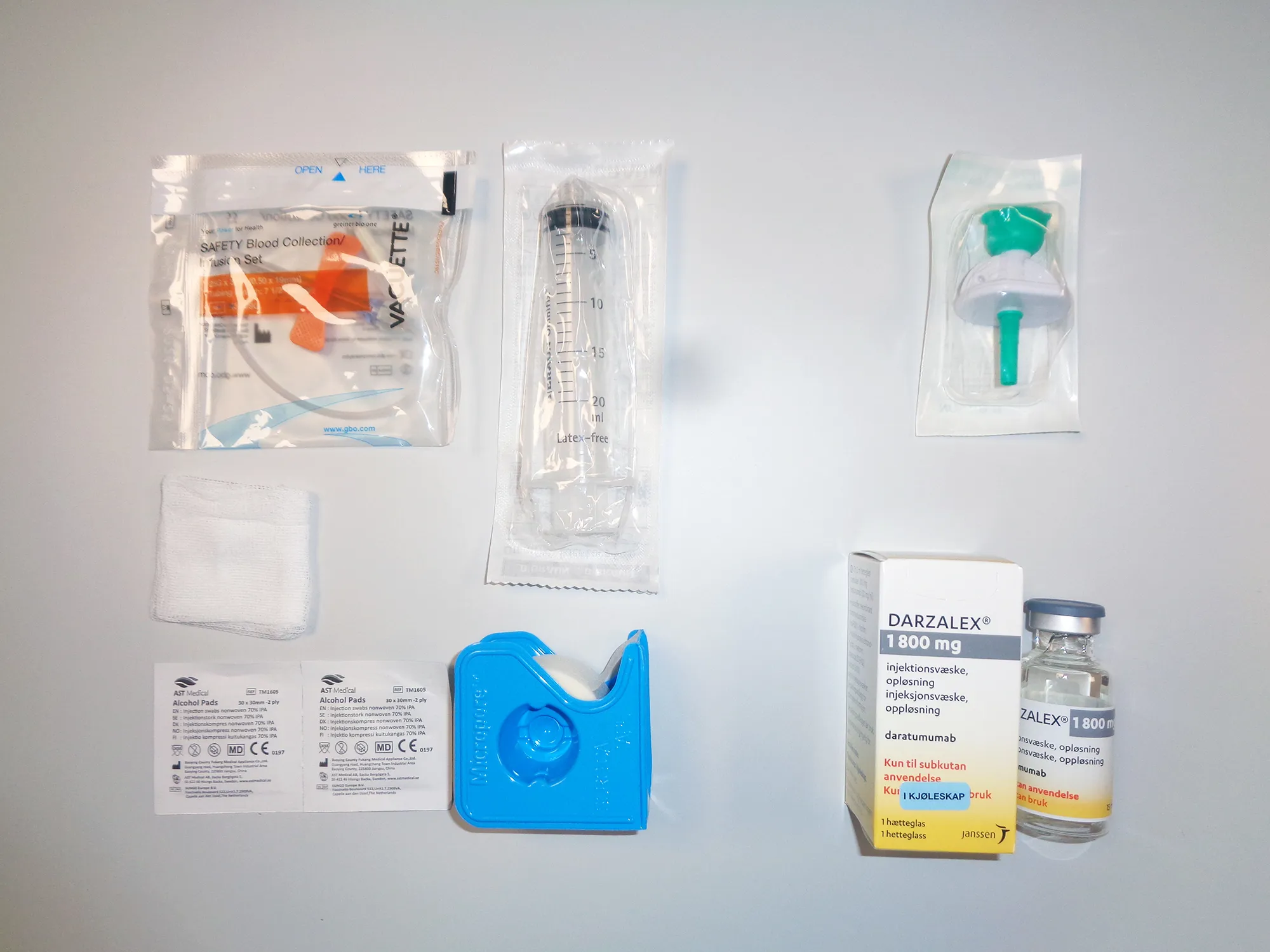 Et sett medisinske engangsartikler og utstyr lagt utover et bord. Øverst ligger en blodprøvenål i emballasje, en plastinnpakket sprøyte og en grønn kanyle i emballasje. Nederst ligger to kompresser, to små etiketter, en blå tapeholder, samt en eske og hetteglass med legemiddelet Darzalex 1800 mg.
