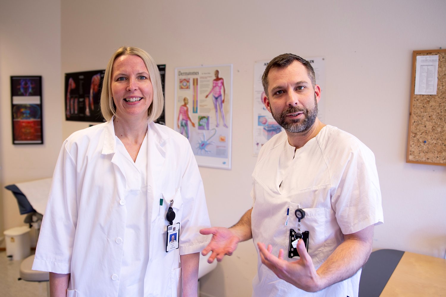 To helsearbeidere i hvite uniformer står i et undersøkelsesrom. Bak dem henger anatomiske plakater og et oppslagstavle. Et behandlingsbord og en krakk er synlige i rommet