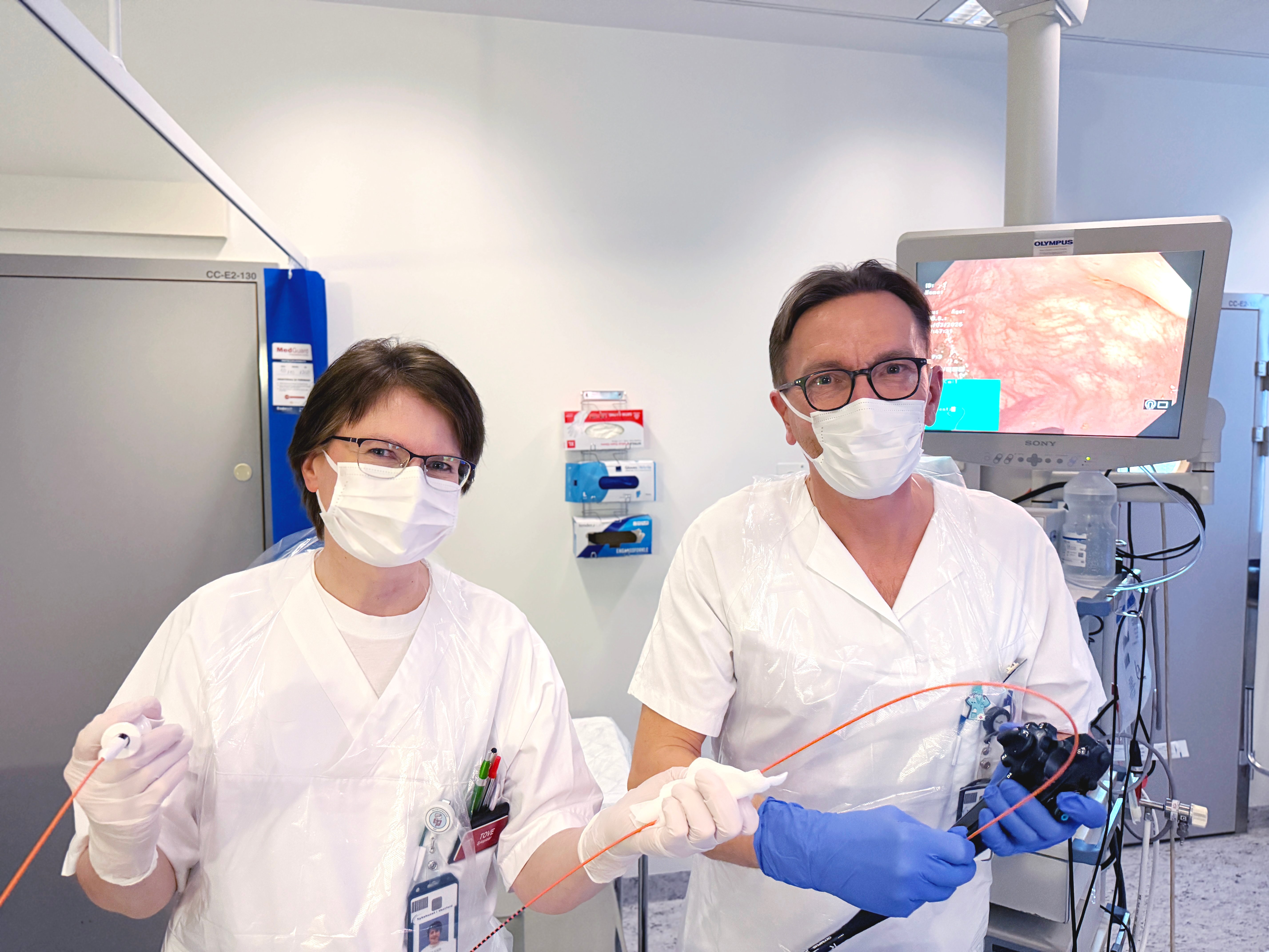 To helsearbeidere står i et undersøkelsesrom og holder medisinsk utstyr for endoskopi. En skjerm i bakgrunnen viser et bilde fra undersøkelsen, og rommet er utstyrt med medisinsk teknisk utstyr og oppheng for forbruksartikler.