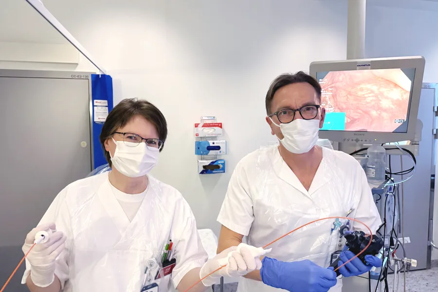 To helsearbeidere står i et undersøkelsesrom og holder medisinsk utstyr for endoskopi. En skjerm i bakgrunnen viser et bilde fra undersøkelsen, og rommet er utstyrt med medisinsk teknisk utstyr og oppheng for forbruksartikler.