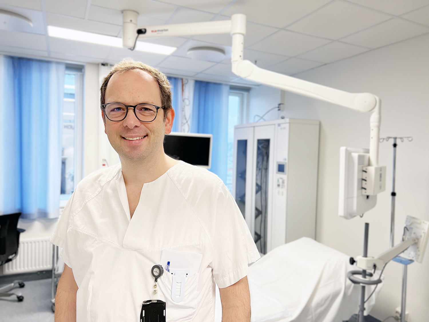 En person i hvitt klinisk arbeidstøy står i et lyst undersøkelsesrom. I bakgrunnen ses medisinsk utstyr, blant annet en behandlingsbenk, monitor, opphengsarm og skap for utstyr.