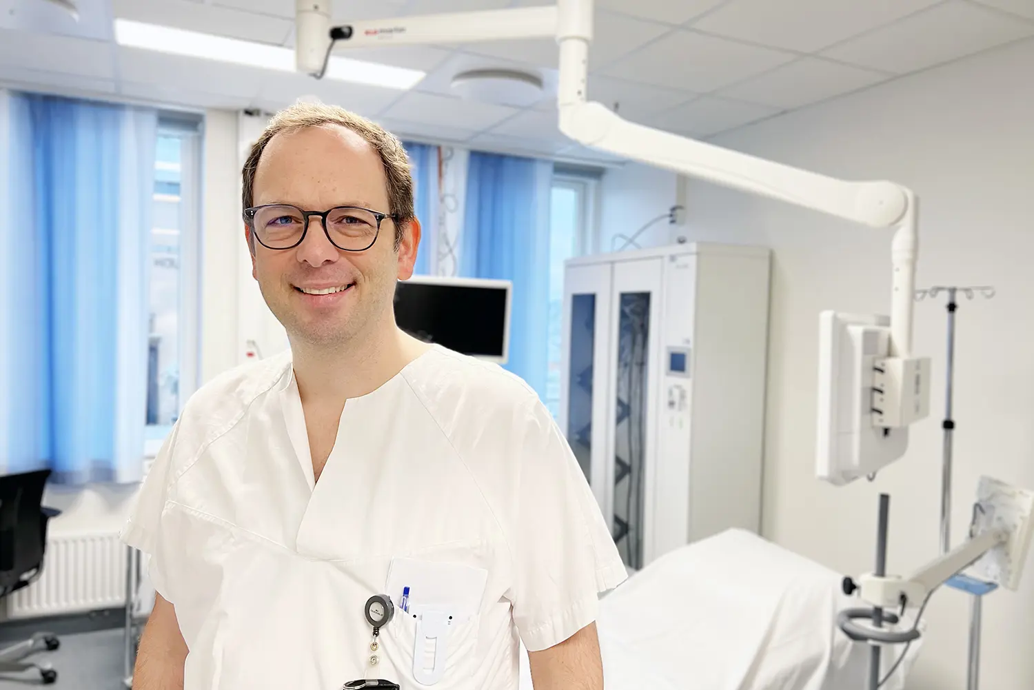 En person i hvitt klinisk arbeidstøy står i et lyst undersøkelsesrom. I bakgrunnen ses medisinsk utstyr, blant annet en behandlingsbenk, monitor, opphengsarm og skap for utstyr.