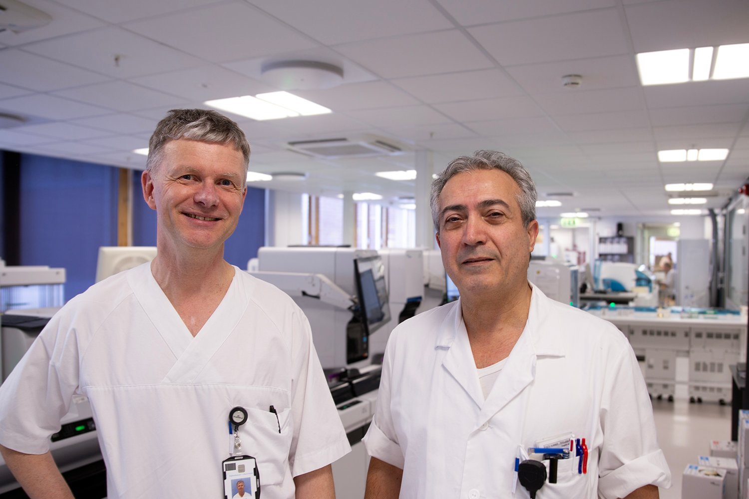 To menn i hvite medisinske uniformer står i et lyst laboratoriemiljø. Den ene har et synlig ID-kort festet til brystlommen. Bak dem står dataskjermer, laboratorieutstyr og arbeidsstasjoner i et ryddig og moderne klinisk rom.