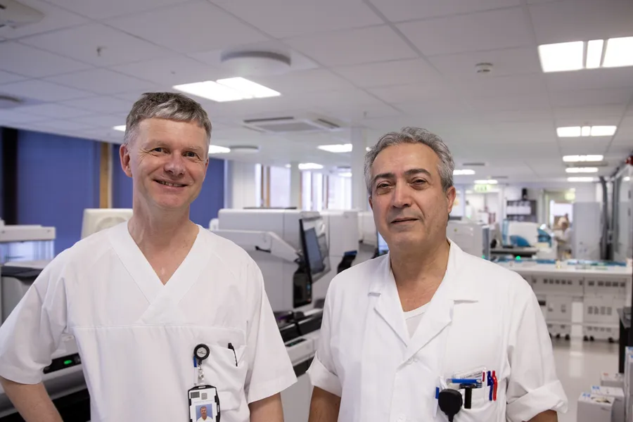 To menn i hvite medisinske uniformer står i et lyst laboratoriemiljø. Den ene har et synlig ID-kort festet til brystlommen. Bak dem står dataskjermer, laboratorieutstyr og arbeidsstasjoner i et ryddig og moderne klinisk rom.