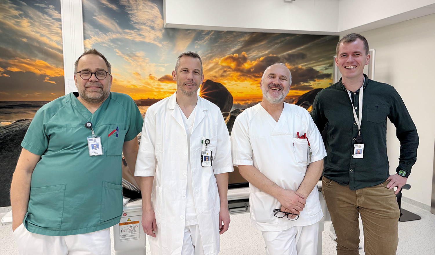 Fire ansatte står sammen i et klinisk miljø. Tre av dem er helsepersonell i hvite frakker, og én er kledd i profesjonelt, uformelt tøy med ID‑kort. Bak dem henger et stort bilde av en solnedgang over en steinete strand. Gruppen smiler mot kameraet.