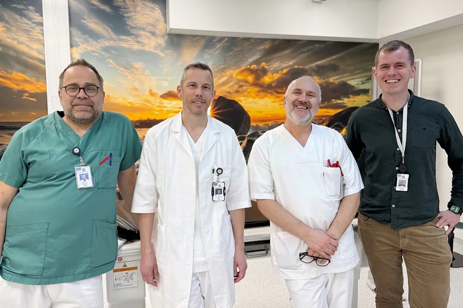 Fire ansatte står sammen i et klinisk miljø. Tre av dem er helsepersonell i hvite frakker, og én er kledd i profesjonelt, uformelt tøy med ID‑kort. Bak dem henger et stort bilde av en solnedgang over en steinete strand. Gruppen smiler mot kameraet.