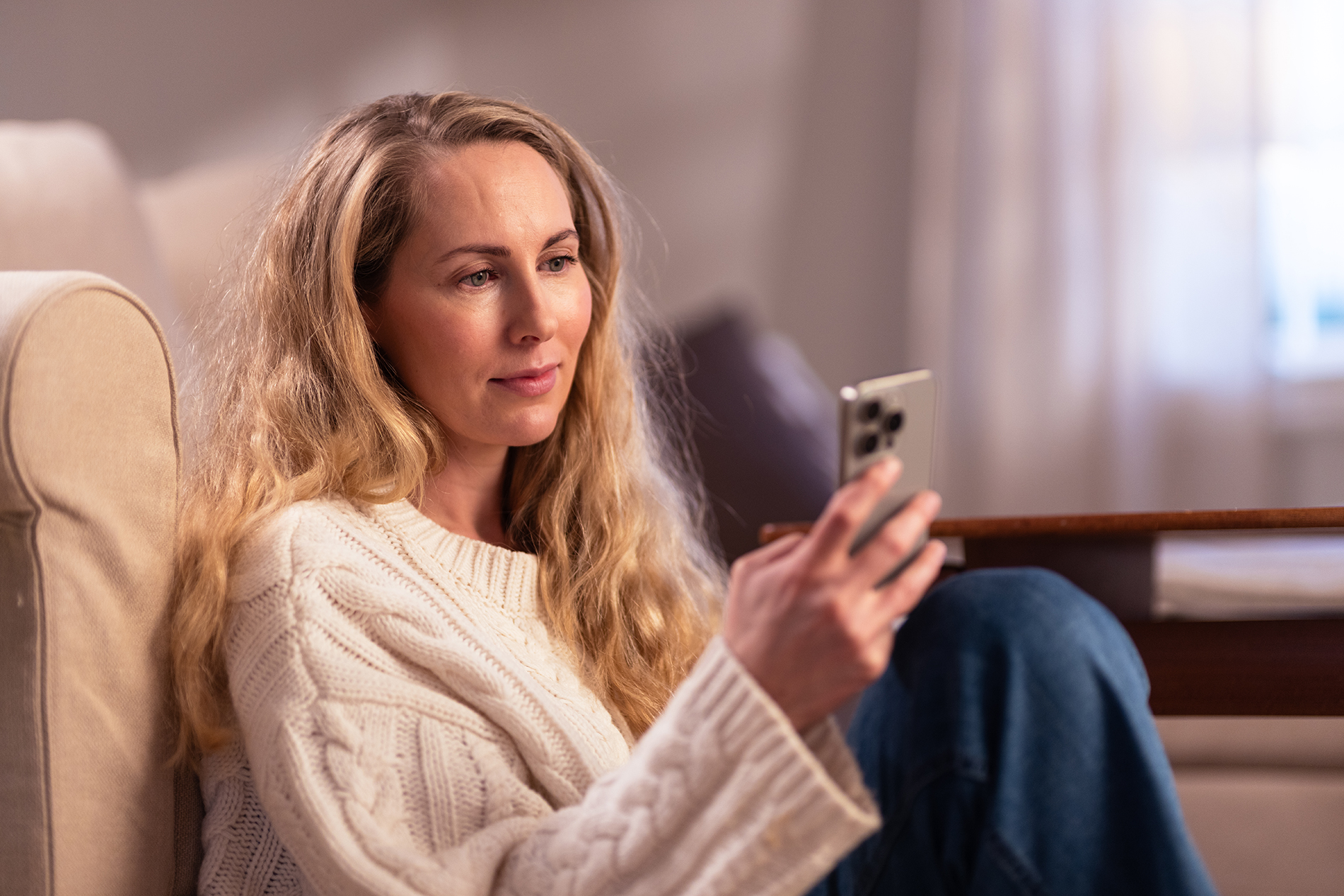 En person sitter innendørs på et møblert oppholdsrom og holder en mobiltelefon. Personen sitter ved siden av en lys sofa, iført en lys strikkegenser og jeans. Interiøret er dempet med myke tekstiler og varmt lys.