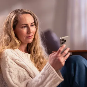 En person sitter innendørs på et møblert oppholdsrom og holder en mobiltelefon. Personen sitter ved siden av en lys sofa, iført en lys strikkegenser og jeans. Interiøret er dempet med myke tekstiler og varmt lys.