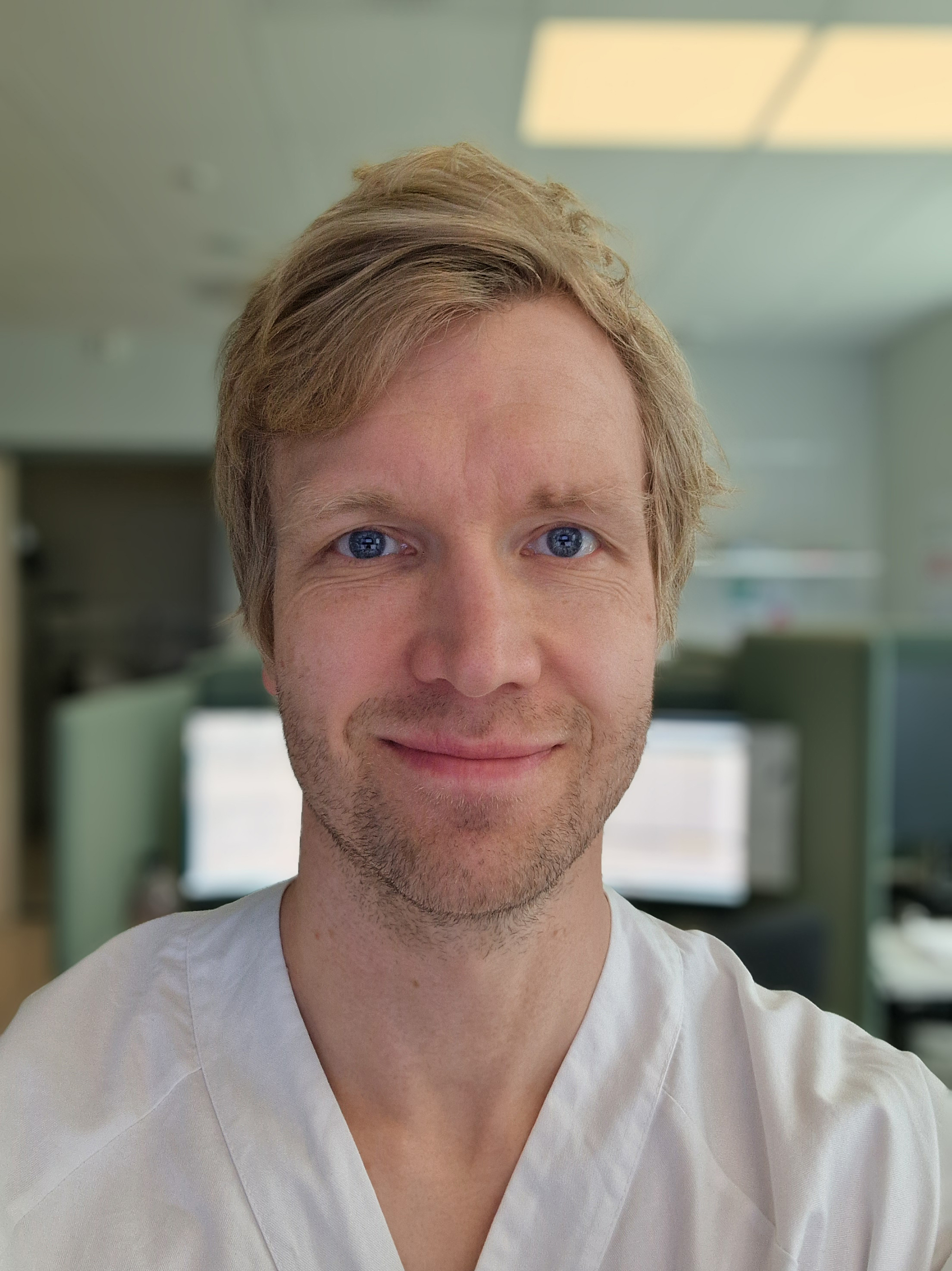 En person i en arbeidsuniform befinner seg innendørs i et rom med kontorutstyr og dataskjermer i bakgrunnen. Personen står nær kamera og ser rett frem.