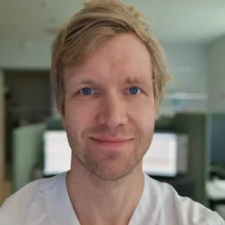 En person i en arbeidsuniform befinner seg innendørs i et rom med kontorutstyr og dataskjermer i bakgrunnen. Personen står nær kamera og ser rett frem.