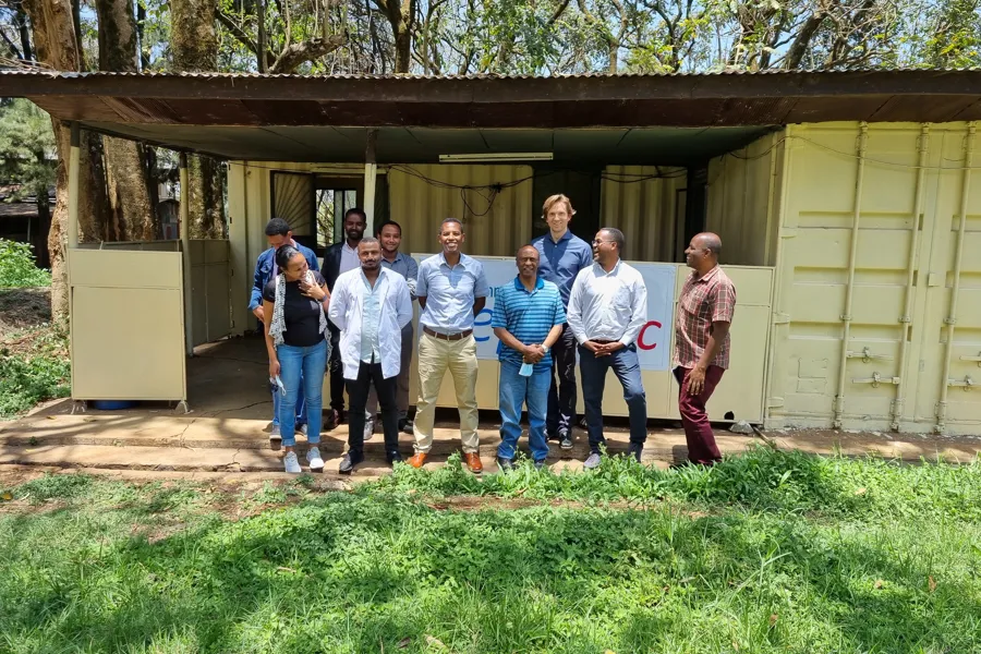 En gruppe mennesker står ute foran en lav bygning med åpne containere eller moduler bak seg. Bygningen ligger i et grønt område med trær i bakgrunnen. Gruppen står i gresset foran strukturen, og bildet er tatt i sollys