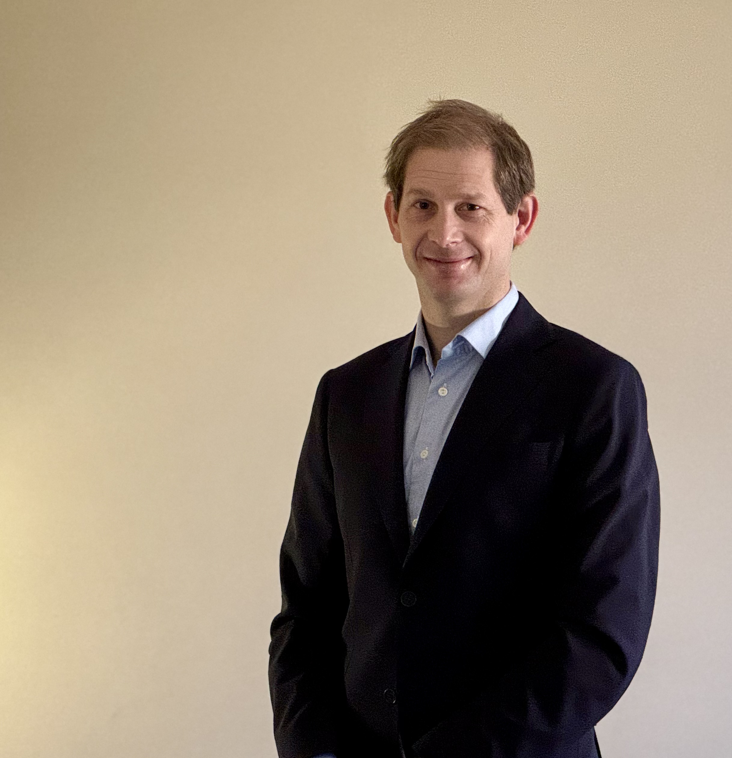 En person står innendørs foran en ensfarget vegg. Personen har kort hår og bruker en mørk jakke over en skjorte. Personen står vendt mot kamera med armene avslappet langs siden