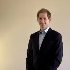 En person står innendørs foran en ensfarget vegg. Personen har kort hår og bruker en mørk jakke over en skjorte. Personen står vendt mot kamera med armene avslappet langs siden