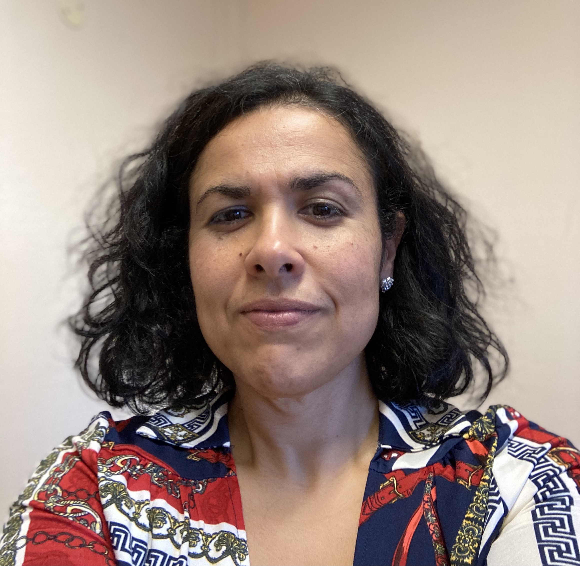 Portrettfoto av en voksen person med skulderlangt krøllet hår, ikledd mønstret skjorte, fotografert mot en lys, nøytral bakgrunn.