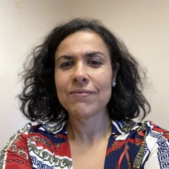 Portrettfoto av en voksen person med skulderlangt krøllet hår, ikledd mønstret skjorte, fotografert mot en lys, nøytral bakgrunn.