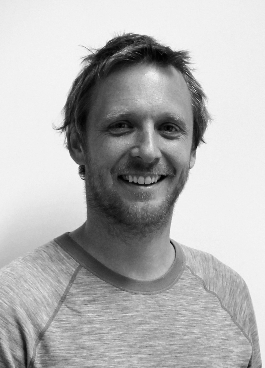 Portrettfoto i svart‑hvitt av en voksen person med kort hår og skjegg, iført ensfarget genser, fotografert mot en lys, nøytral bakgrunn.