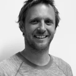 Portrettfoto i svart‑hvitt av en voksen person med kort hår og skjegg, iført ensfarget genser, fotografert mot en lys, nøytral bakgrunn.