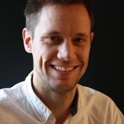 Portrettfoto av en voksen person med kort hår, iført lys skjorte, fotografert mot en mørk, nøytral bakgrunn.