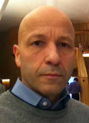 Portrettfoto av en voksen person med kort hår, iført skjorte og genser, fotografert innendørs mot en bakgrunn med varmt trepreg.