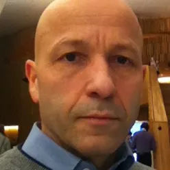 Portrettfoto av en voksen person med kort hår, iført skjorte og genser, fotografert innendørs mot en bakgrunn med varmt trepreg.