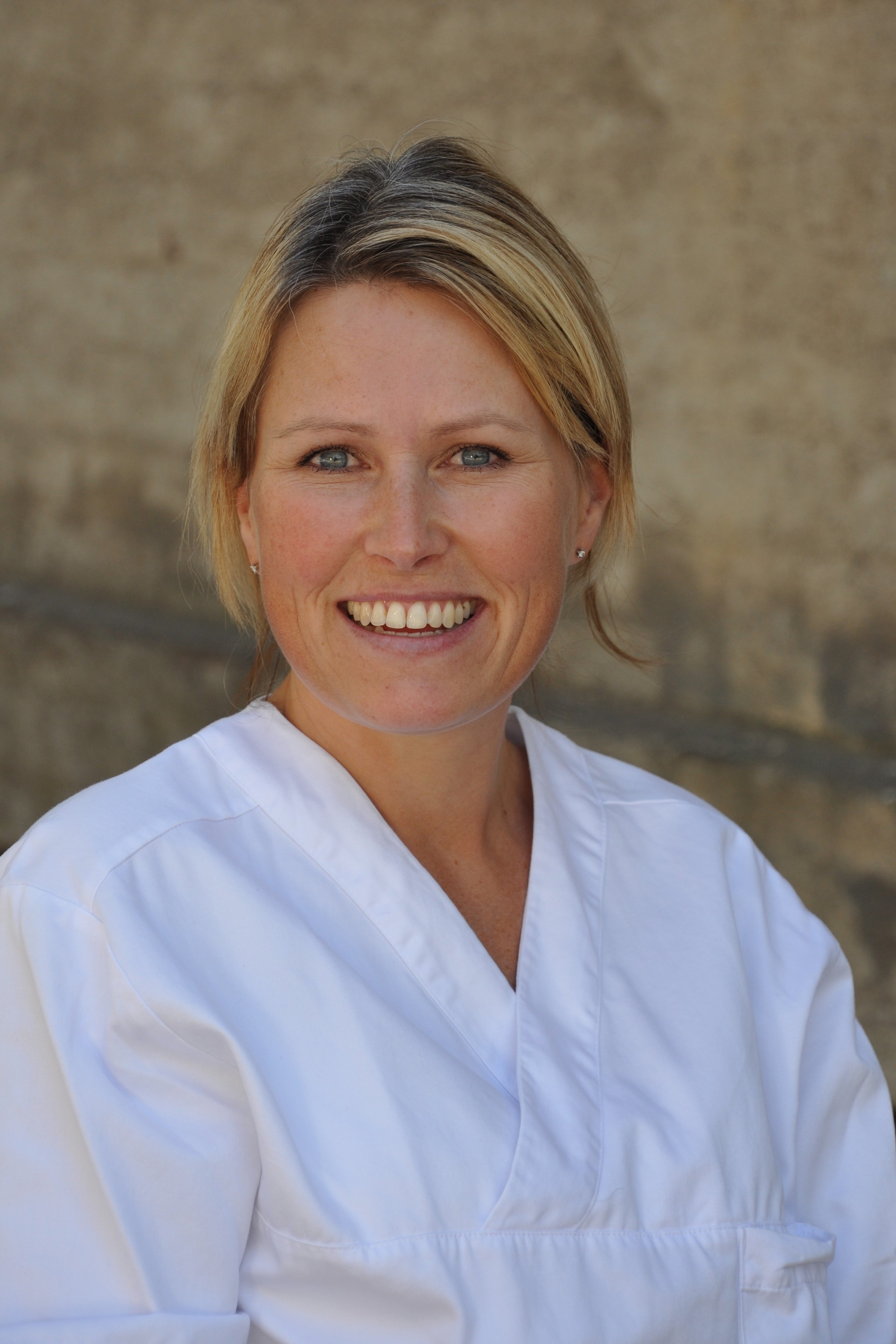 Portrettfoto av en voksen person med kort, lyst hår, iført hvit overdel, fotografert utendørs mot en beige steinbakgrunn.