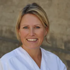 Portrettfoto av en voksen person med kort, lyst hår, iført hvit overdel, fotografert utendørs mot en beige steinbakgrunn.