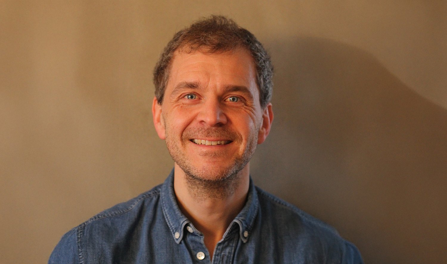 En mann står vendt mot kamera foran en ensfarget beige bakgrunn. Personen har kort, lysebrunt hår og bruker en blå denimskjorte. Lyset er varmt og jevnt og gjør ansiktstrekkene tydelige.