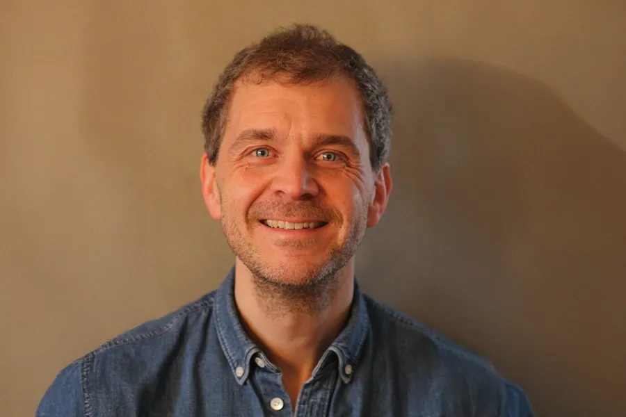 En mann står vendt mot kamera foran en ensfarget beige bakgrunn. Personen har kort, lysebrunt hår og bruker en blå denimskjorte. Lyset er varmt og jevnt og gjør ansiktstrekkene tydelige.