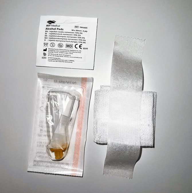 Sterilt bandasjeutstyr bestående av alkoholkserviett, venekanyle i emballasje og kompress med teip, lagt utover på hvit bakgrunn.