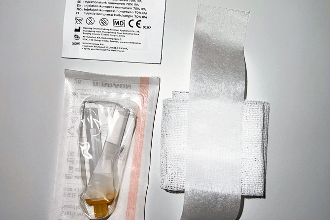 Sterilt bandasjeutstyr bestående av alkoholkserviett, venekanyle i emballasje og kompress med teip, lagt utover på hvit bakgrunn.
