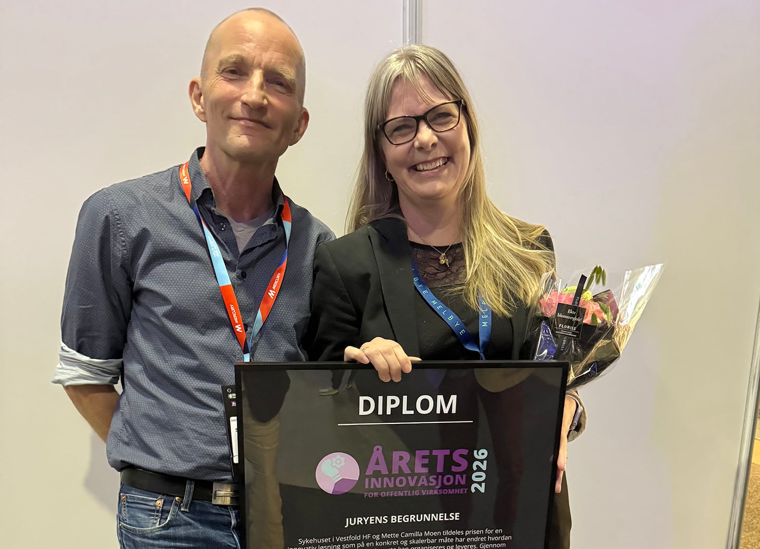 To personer står side om side på en prisutdeling. Den ene holder en blomsterbukett og et innrammet diplom merket «Årets innovasjon 2026 for offentlig virksomhet». Begge har navneskilt rundt halsen og smiler mot kameraet foran en nøytral bakgrunn.
