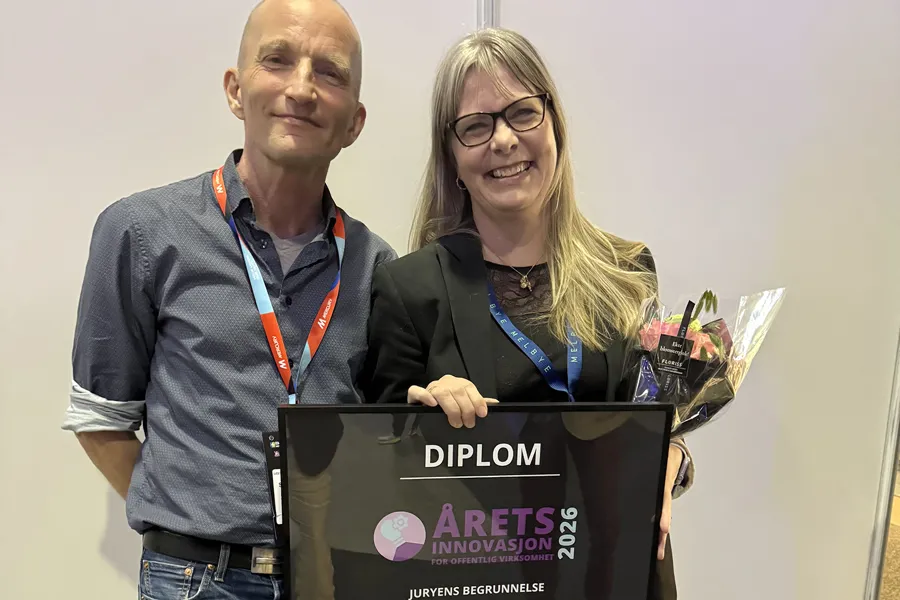 To personer står side om side på en prisutdeling. Den ene holder en blomsterbukett og et innrammet diplom merket «Årets innovasjon 2026 for offentlig virksomhet». Begge har navneskilt rundt halsen og smiler mot kameraet foran en nøytral bakgrunn.