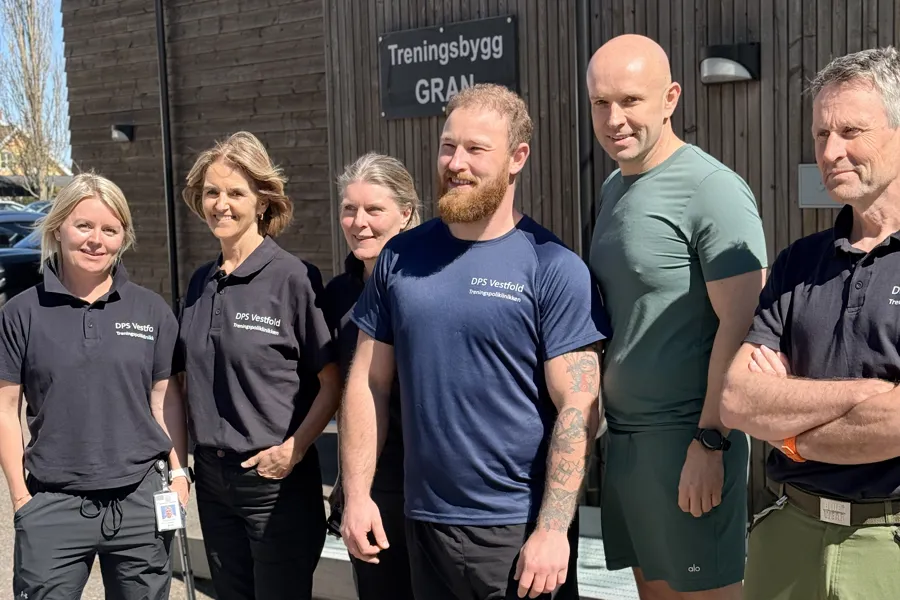 En gruppe på seks ansatte står ute foran bygningen «Treningsbygg GRAN». Flere av dem har på seg mørke T‑skjorter med teksten «DPS Vestfold Treningspoliklinikken». De smiler mot kameraet i sollys.