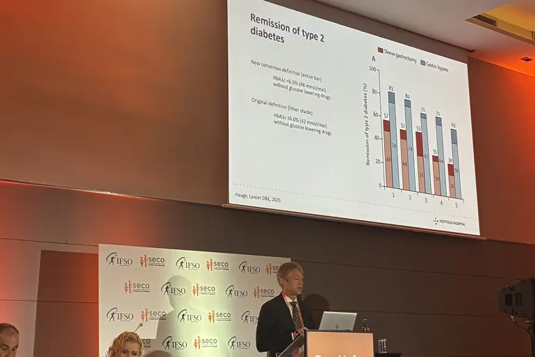 En foredragsholder står på en konferansescene og presenterer en slide med grafer som viser remisjon av type 2‑diabetes etter ulike typer fedmekirurgi. Bakgrunnen viser konferanselogoer.