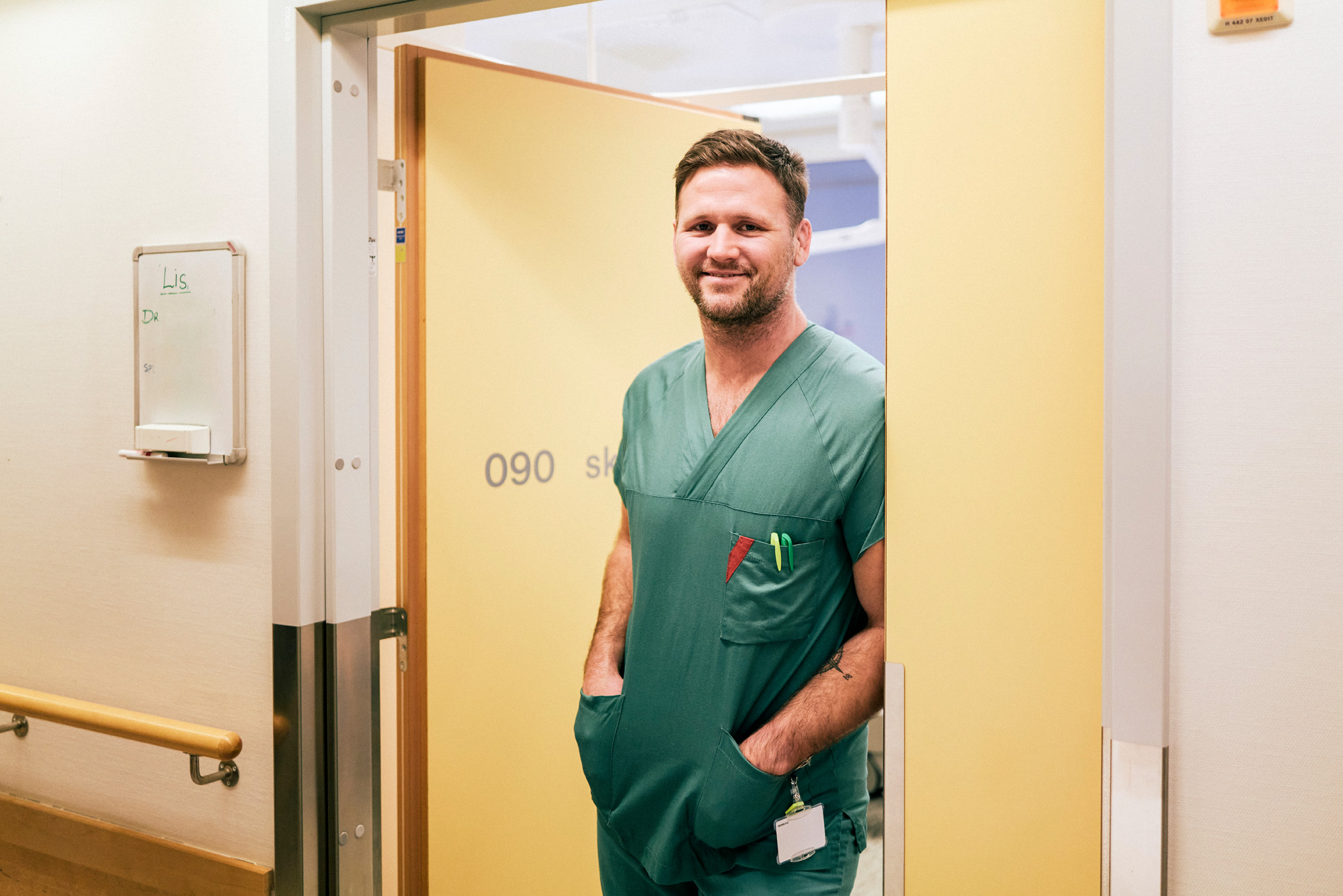 En mann i grønne arbeidsklær står i en døråpning og smiler
