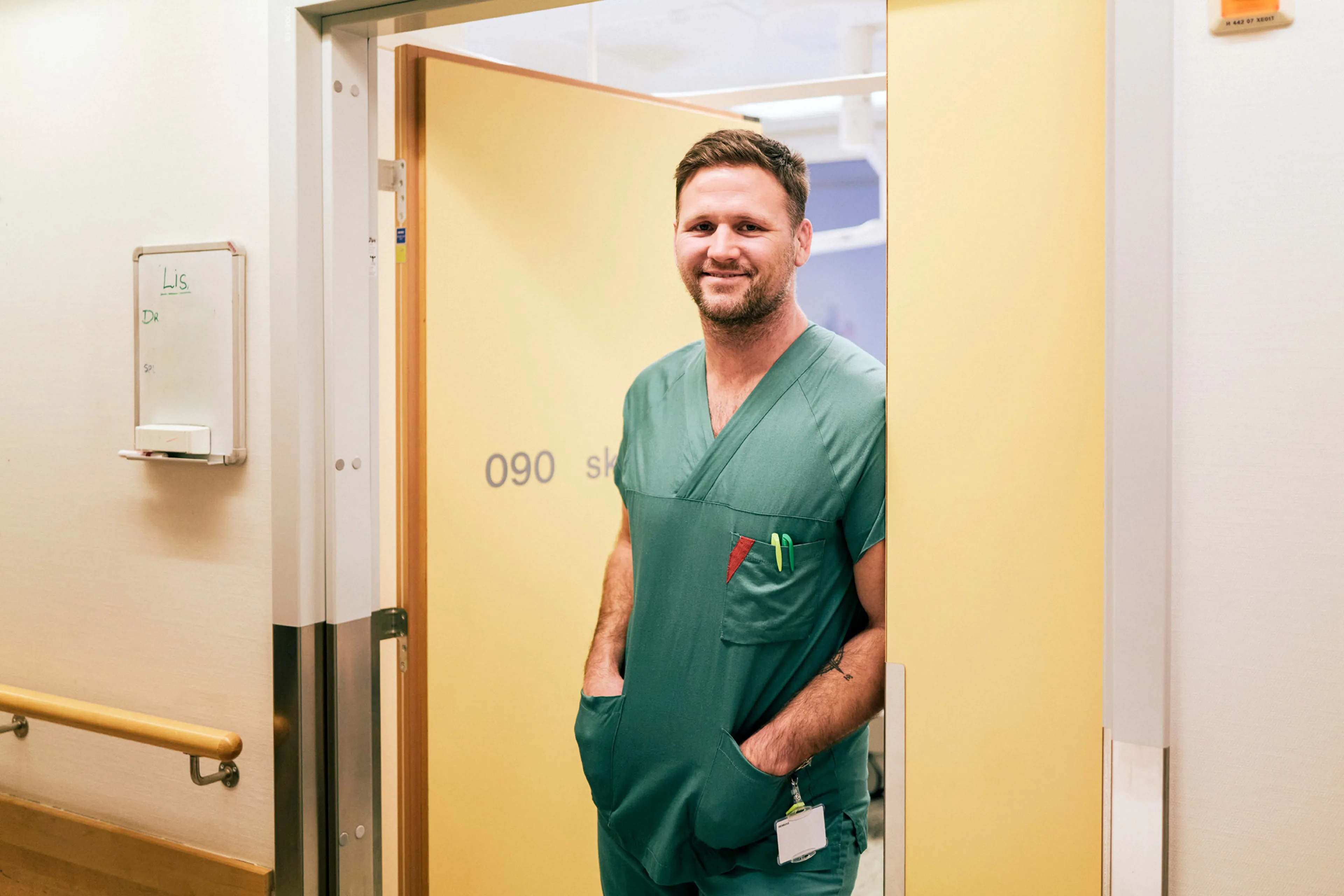 En mann i grønne arbeidsklær står i en døråpning og smiler