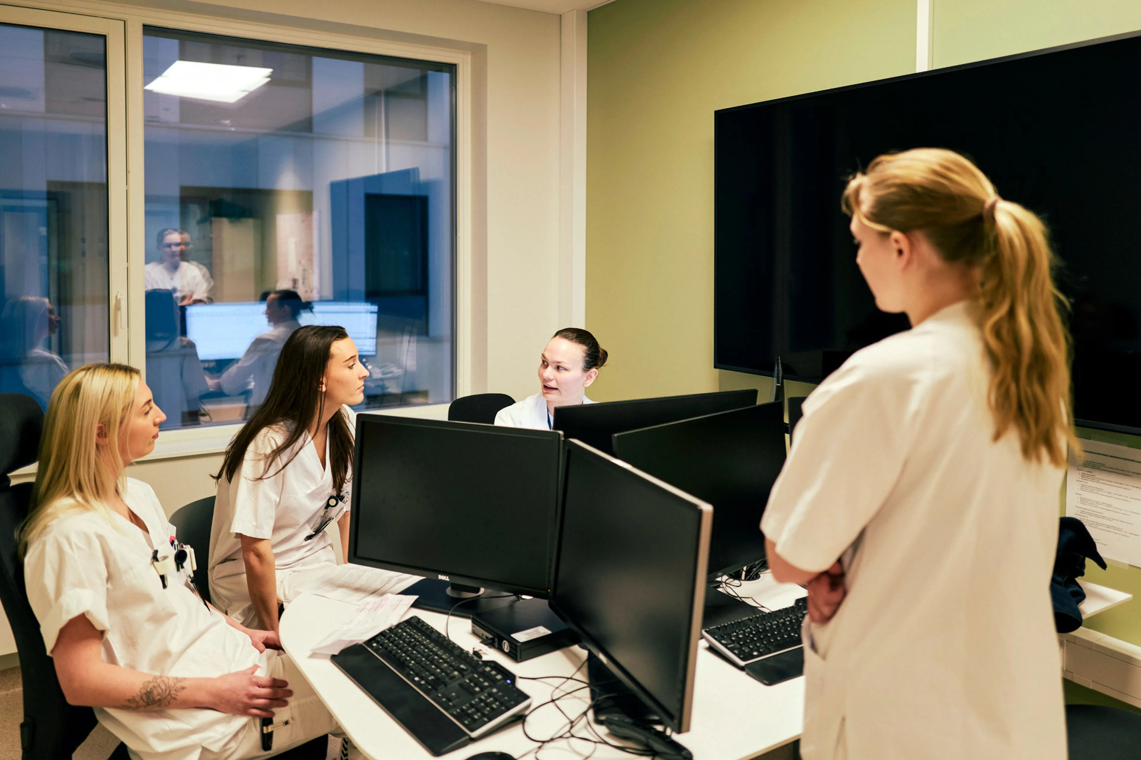Helsepersonell i hvitt arbeidsantrekk prater sammen på et kontor med mange dataskjermer