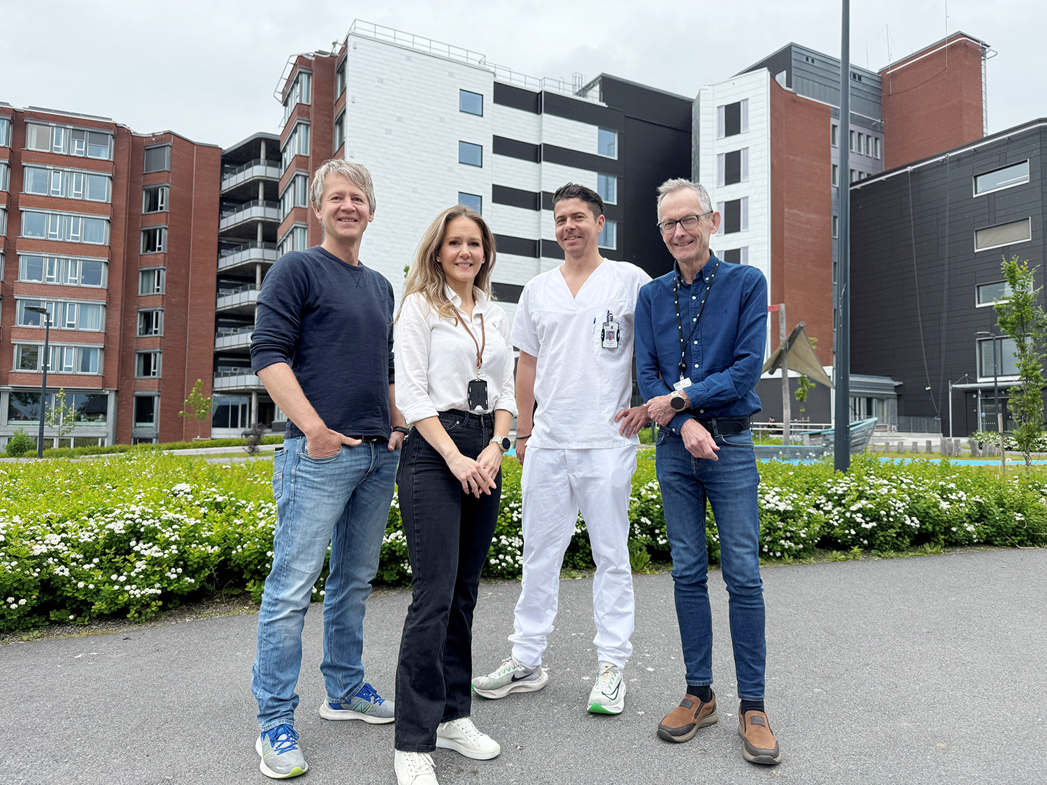 En gruppe på fire personer står ute foran Sykehuset i Vestfold, et moderne bygg med fasader i murstein og lyse paneler. En av dem er kledd i hvitt sykehusantrekk, mens de andre har hverdags- eller businessklær. De står på en steinlagt plass omgitt av gress og hvite blomster, med bolig- eller institusjonsbygg i bakgrunnen under en overskyet himmel.