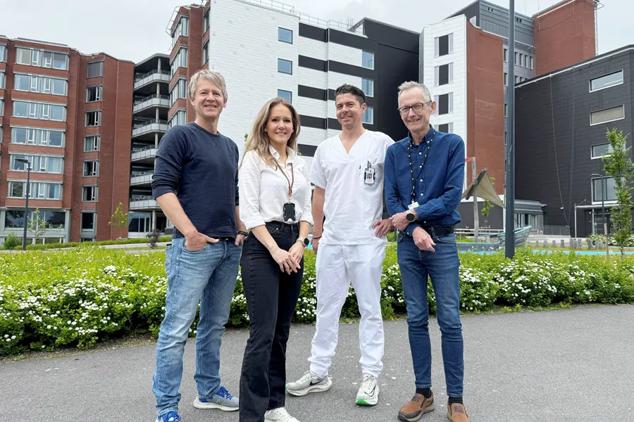 En gruppe på fire personer står ute foran Sykehuset i Vestfold, et moderne bygg med fasader i murstein og lyse paneler. En av dem er kledd i hvitt sykehusantrekk, mens de andre har hverdags- eller businessklær. De står på en steinlagt plass omgitt av gress og hvite blomster, med bolig- eller institusjonsbygg i bakgrunnen under en overskyet himmel.