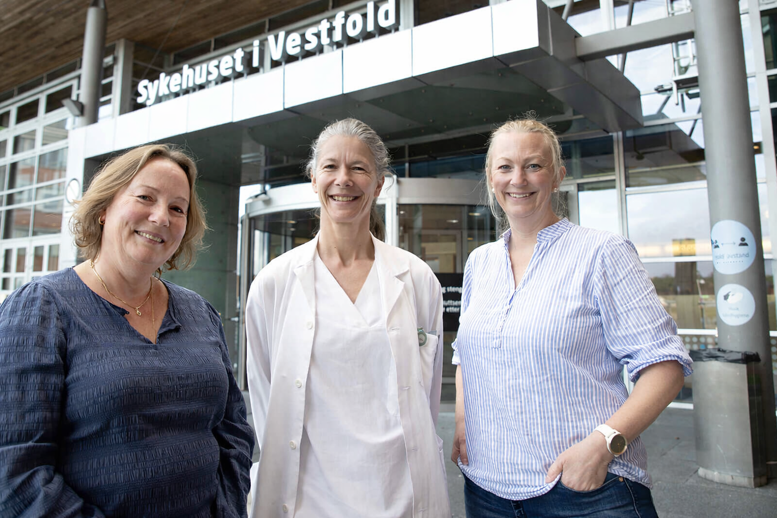 Tre personer står foran hovedinngangen til Sykehuset i Vestfold; en helsearbeider i hvit frakk i midten og to besøkende på hver side.