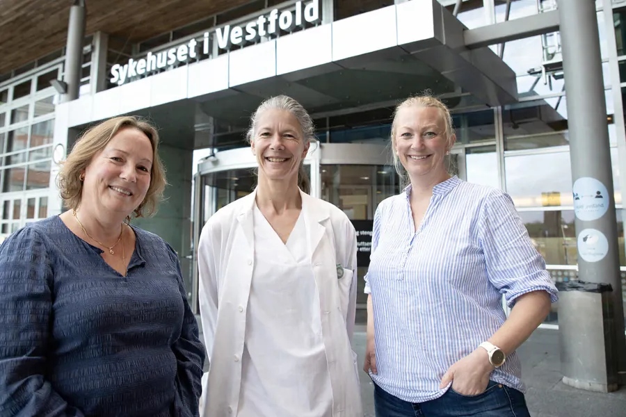 Tre personer står foran hovedinngangen til Sykehuset i Vestfold; en helsearbeider i hvit frakk i midten og to besøkende på hver side.