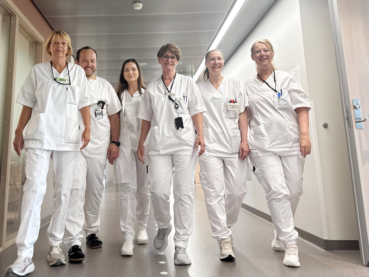 En gruppe på seks helsearbeidere i hvite uniformer går sammen ned en lys sykehuskorridor. De bærer ID‑kort og utstyr i brystlommene, og beveger seg i samlet retning som et team.