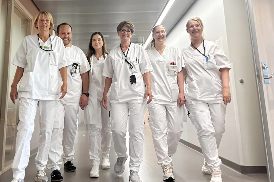 En gruppe på seks helsearbeidere i hvite uniformer går sammen ned en lys sykehuskorridor. De bærer ID‑kort og utstyr i brystlommene, og beveger seg i samlet retning som et team.
