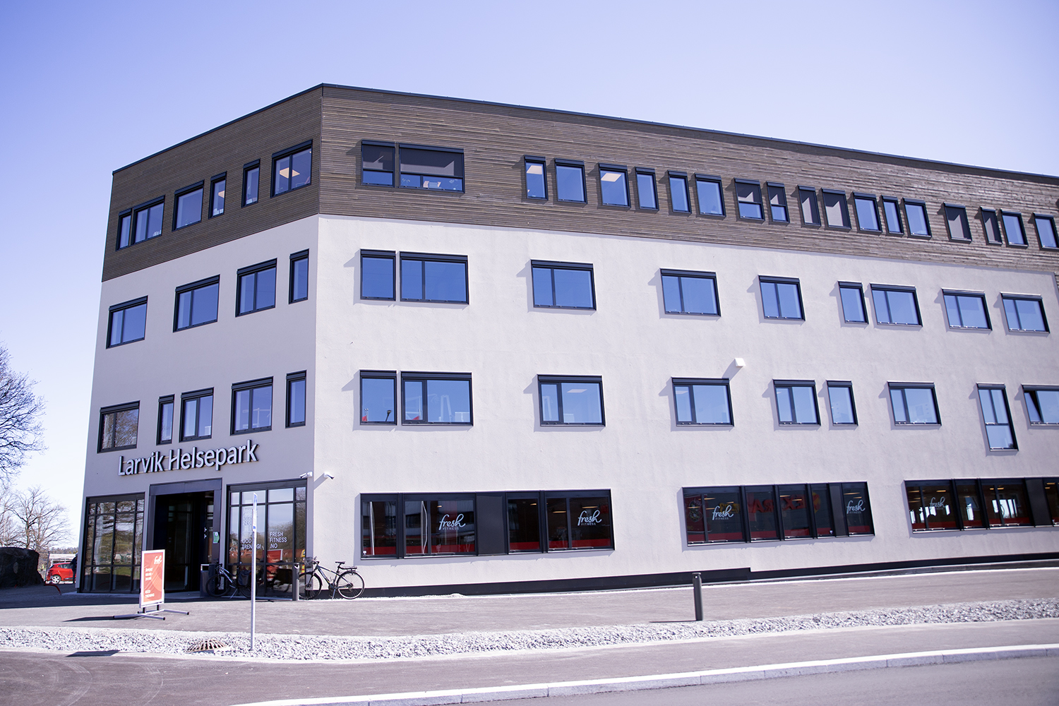 Stort, moderne bygg merket «Larvik Helsepark», sett fra siden og inngangspartiet. Fasaden er lys med en øvre etasje i tre, og mange jevnt plasserte vinduer. Ved inngangen står en sykkel, og gjennom vinduene er treningssenteret «Fresh Fitness» synlig. Asfaltert område og vei i forgrunnen, klart dagslys.