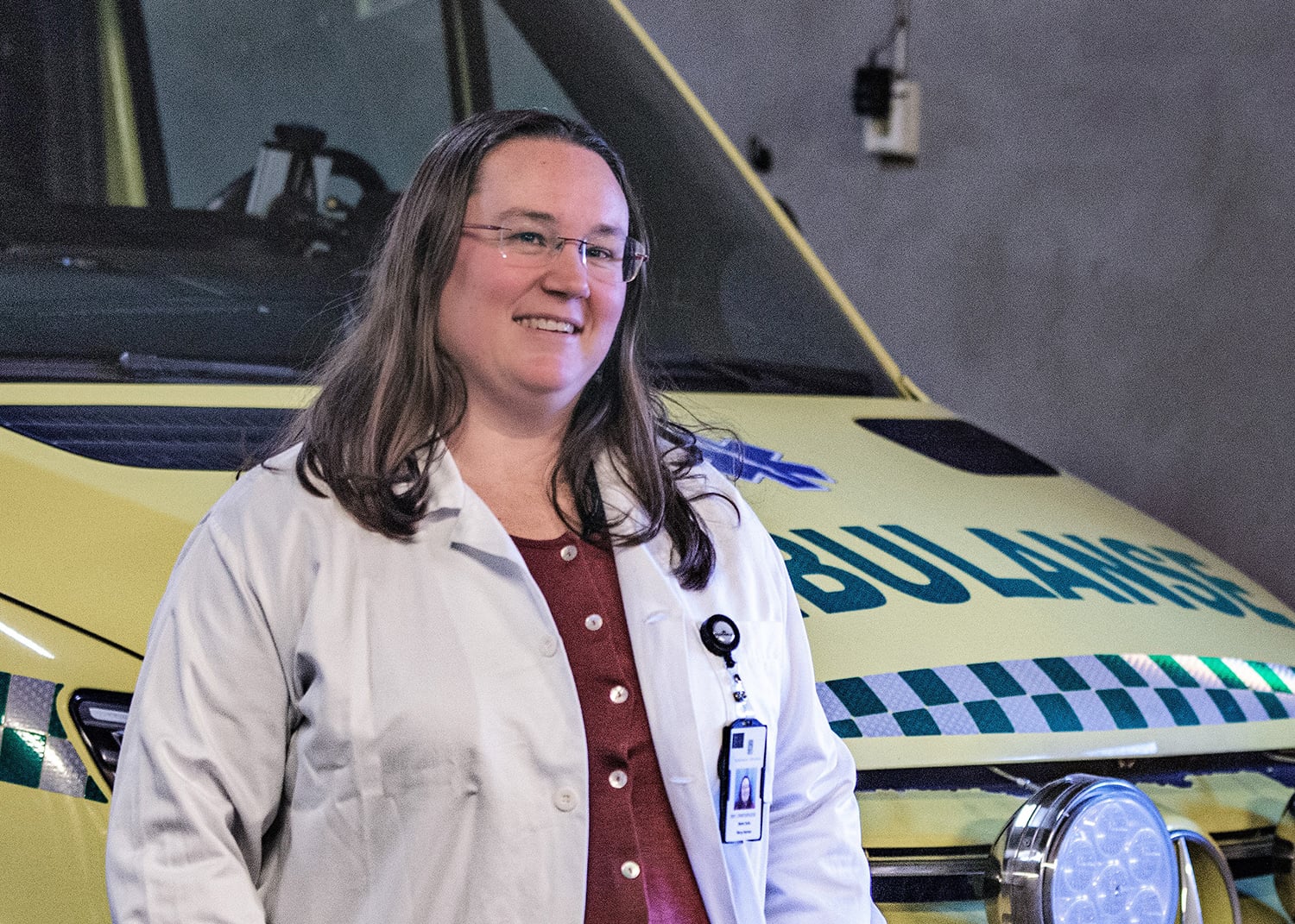 En kvinnelig helsearbeider i hvit frakk og briller står innendørs foran en gul ambulanse med teksten «AMBULANSE» på fronten. Personen har ID‑kort festet til frakken og ser rett mot kamera.