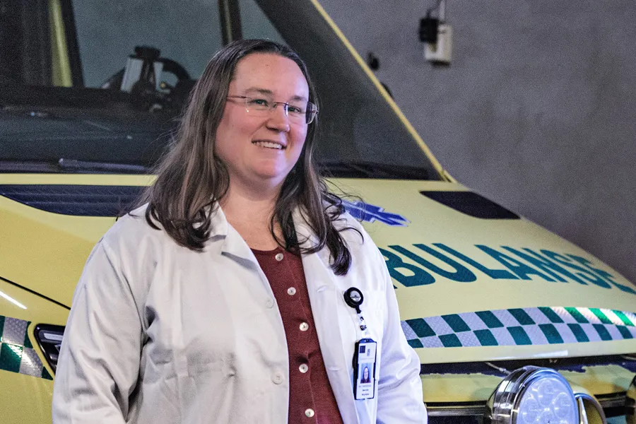 En kvinnelig helsearbeider i hvit frakk og briller står innendørs foran en gul ambulanse med teksten «AMBULANSE» på fronten. Personen har ID‑kort festet til frakken og ser rett mot kamera.
